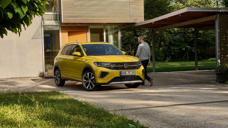 A man walking past a Volkswagen T-Cross, parked outside of a house. The T-Cross is Rubber Ducky yellow.