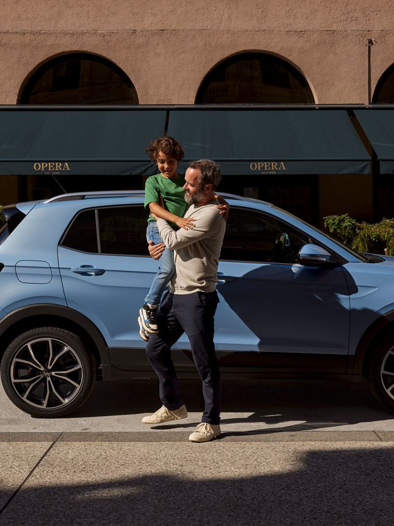 a man carrying a child in front of a blue parked T-Cross