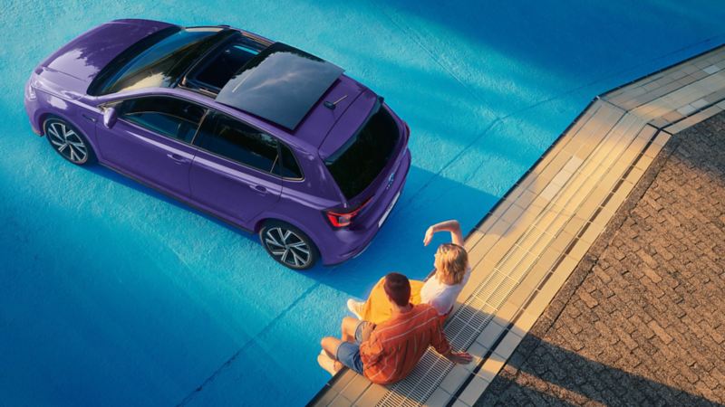 A purple VW Polo with an optional panoramic sliding roof is parked in a hall, a couple is sitting  behind it