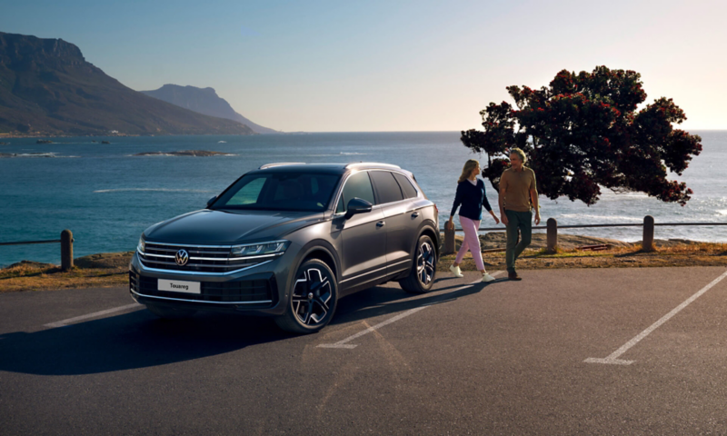 A Touareg seen from the front parked in a car park next to the sea with the owners walking past the car