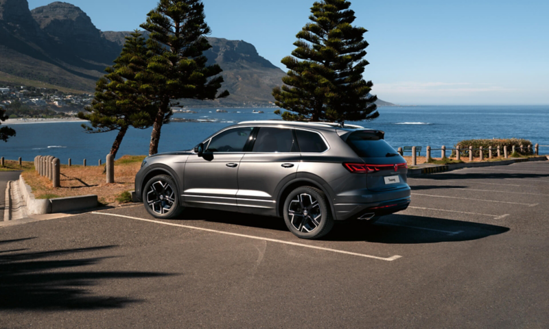 A VW Touareg Elegance parked in the sun, view from the side, behind it you can see the sea and the coast.