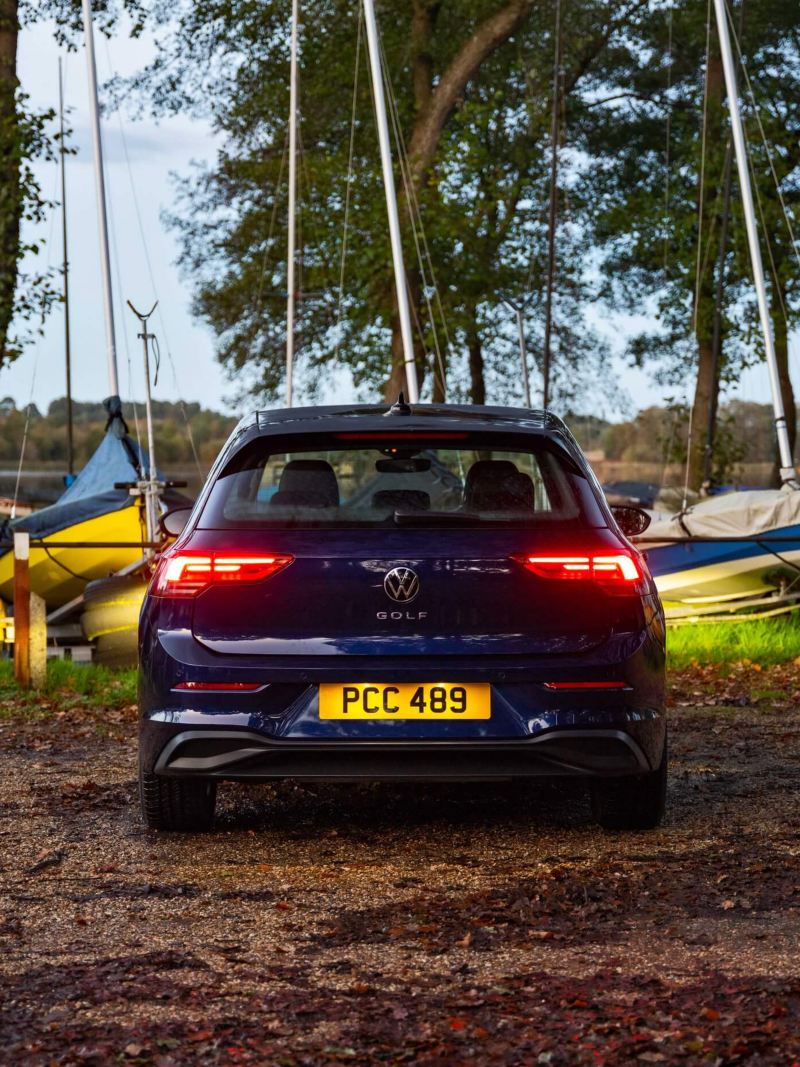 Back of a dark blue new Golf 8 with its break lights on parked in front of some boats at a boat yard.