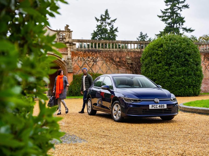 Dark blue Volkswagen Golf 8 parked in a large driveway with 2 people walking away from it smiling and talking