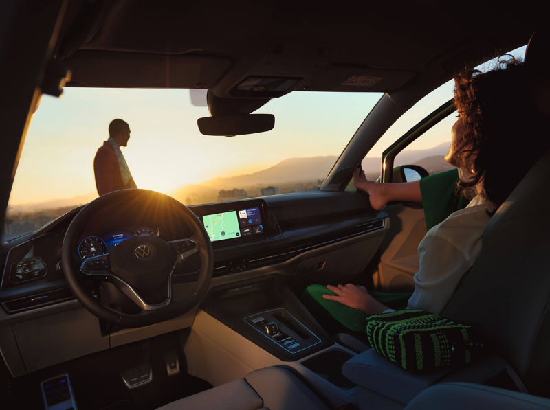 A VW Golf at sunset with a woman in the passenger seat looking at a man standing ahead of the car