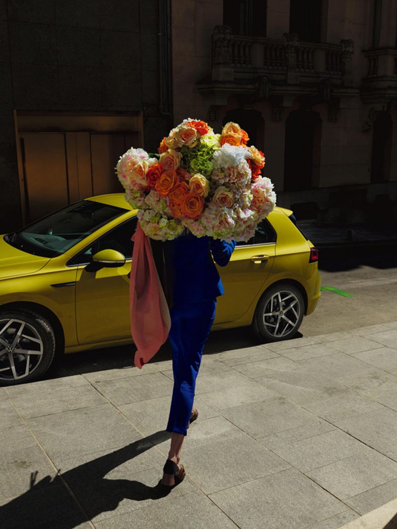 A woman walking with a bunch of flowers past a yellow Golf 8.