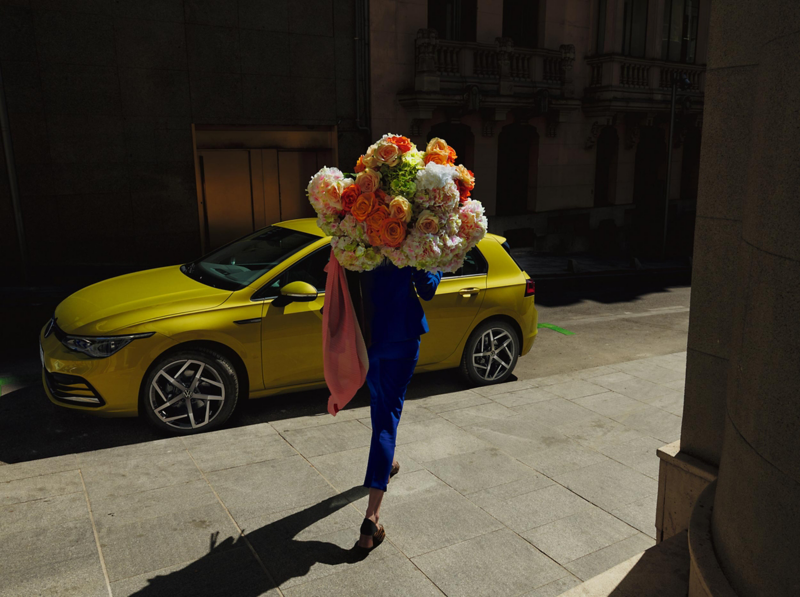 A woman walking with a bunch of flowers past a yellow Golf 8.