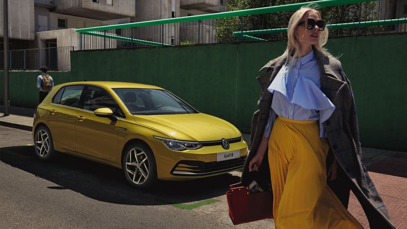 A yellow VW Golf 8 is parked on street with person walking in front of car holding handbag