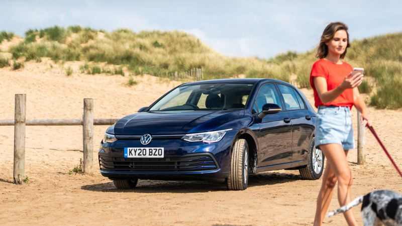 A blue new Golf 8 at the beach and a lady walking past with her dog