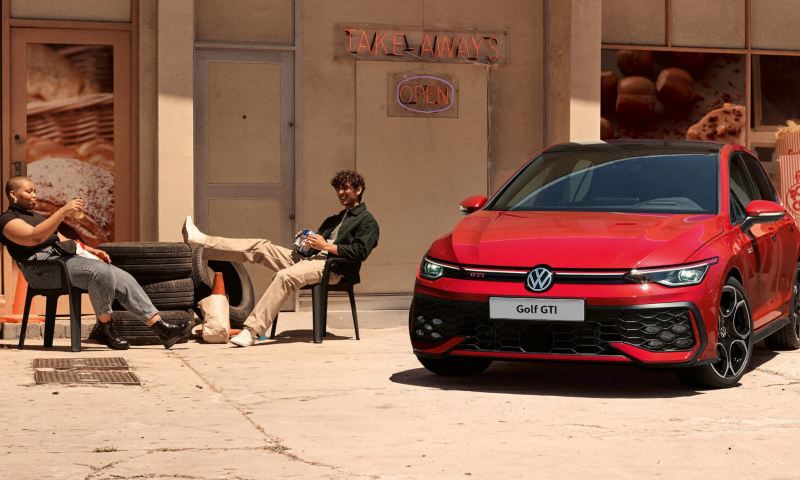 Two people sitting outside a shopfront by a red Golf GTI