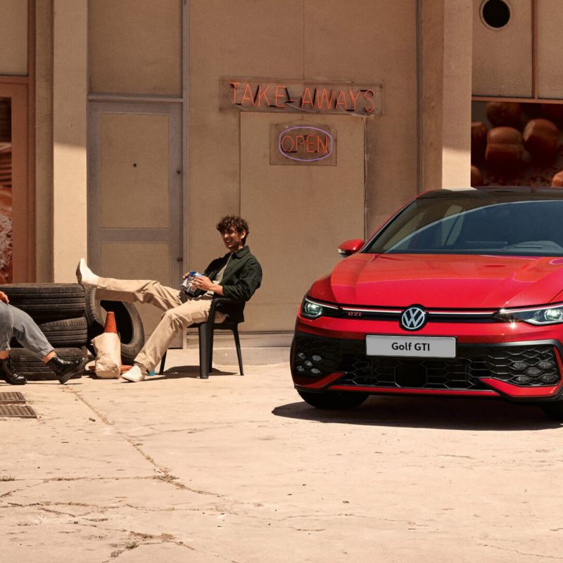 Two people sitting outside a shopfront by a red Golf GTI