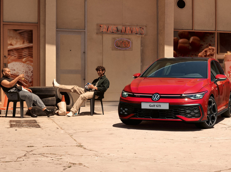Two people sitting outside a shopfront by a red Golf GTI