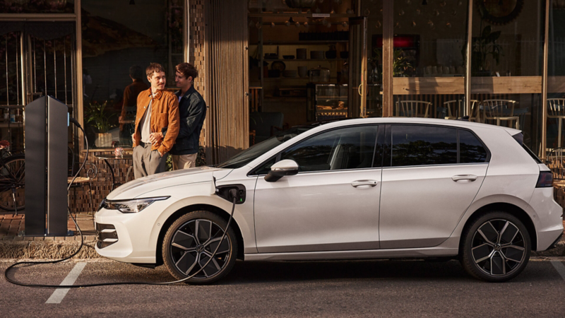 A Golf GTE parked outside of a cafe and plugged into a charging unit with a couple standing beside the car whilst embracing each other