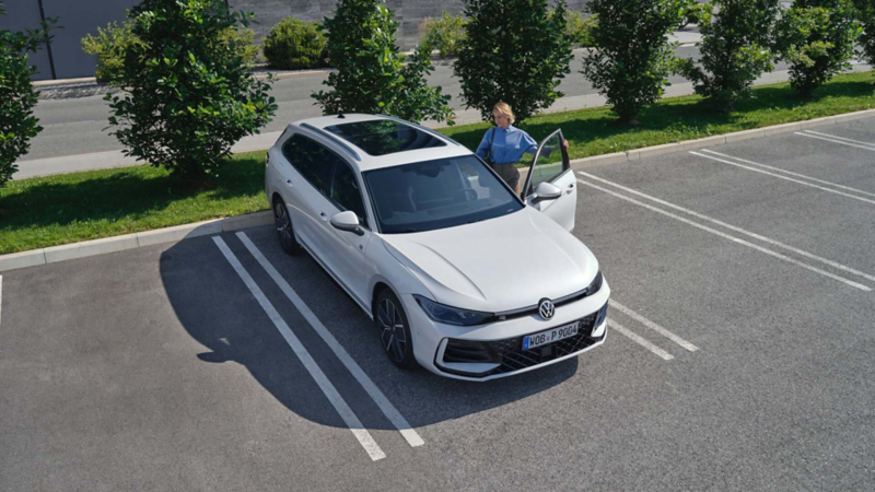 Aerial view of a white Volkswagen Passat car parked in a car park beside bushes. A person in a blue shirt stands by the open driver's door, creating a calm atmosphere.