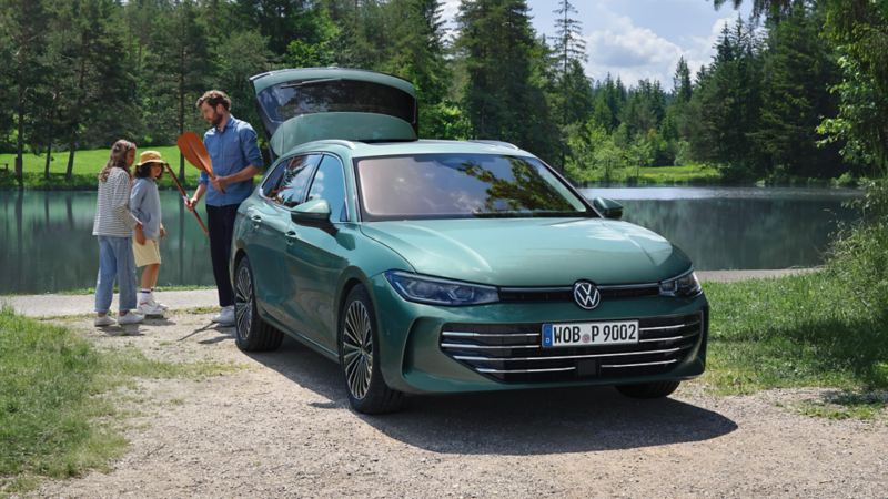 Bluish green VW Passat parked at a swimming lake with the luggage compartment open. An adult hands out recreational toys to two children.