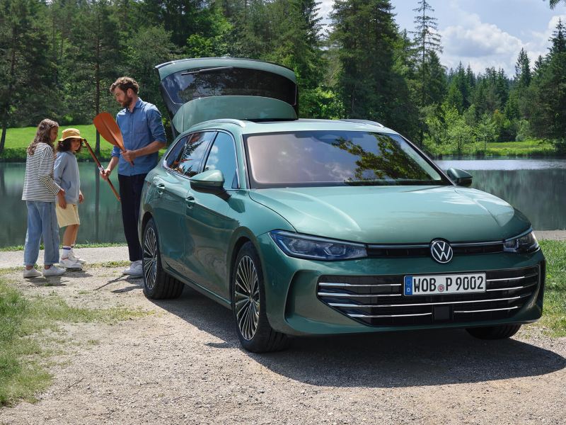 Bluish green VW Passat parked at a swimming lake with the luggage compartment open. An adult hands out recreational toys to two children.