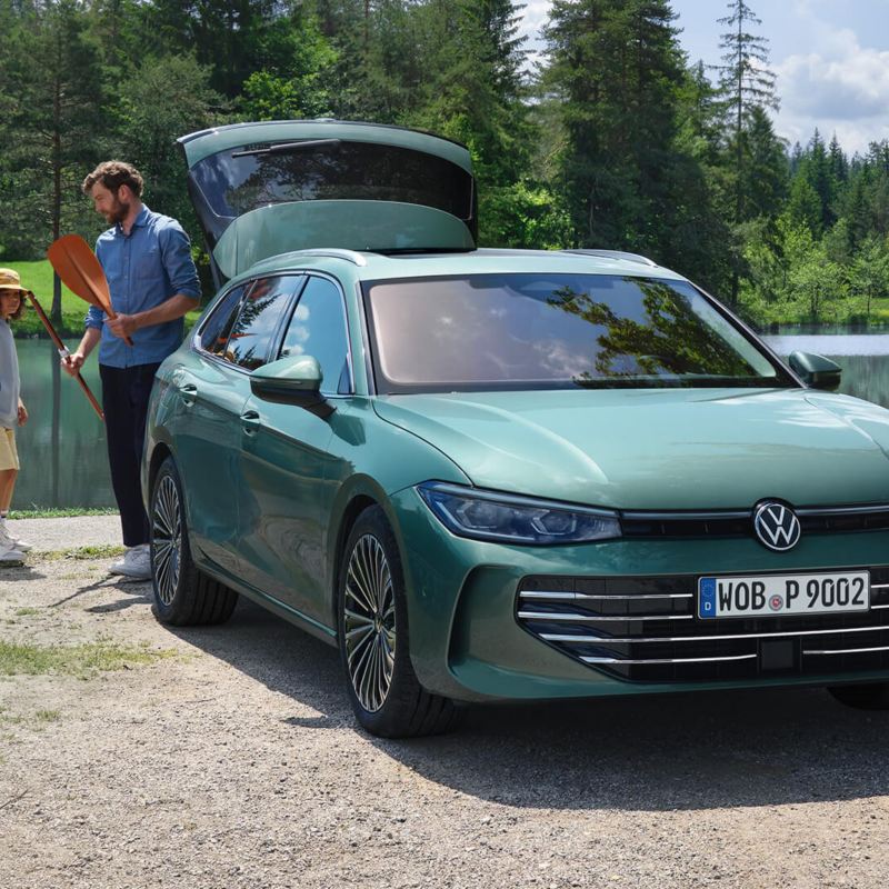 Bluish green VW Passat parked at a swimming lake with the luggage compartment open. An adult hands out recreational toys to two children.