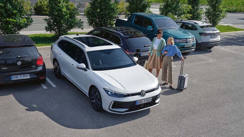 Two people with luggage talking next to a white VW Passat in a car par
