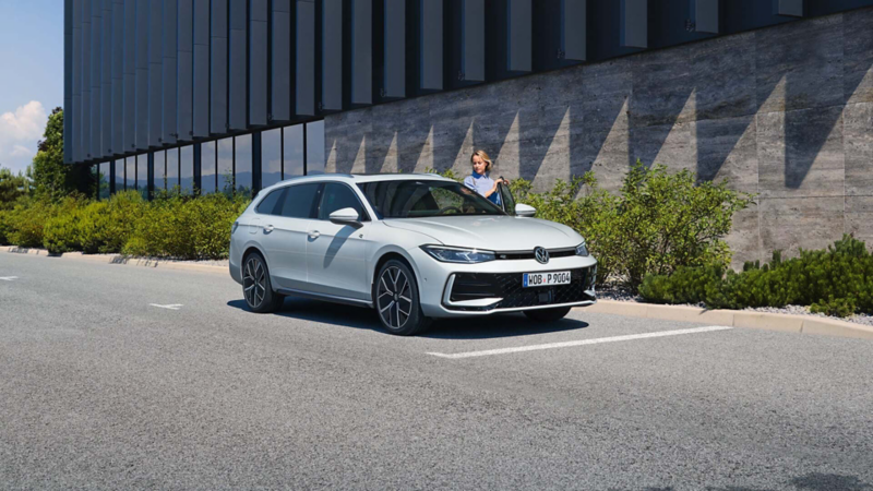 A sleek, modern white Volkswagen Passat driving on a road, showcasing its dynamic design. The background features a blurred urban landscape with greenery.