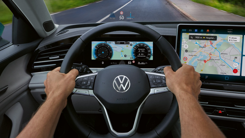 Interior view of VW Passat showing the driver side with large colour display and head-up display. Two hands clasp the steering wheel. 