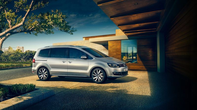 A silver Volkswagen Sharan, parked on the drive of a single story wooden house, trees in the background