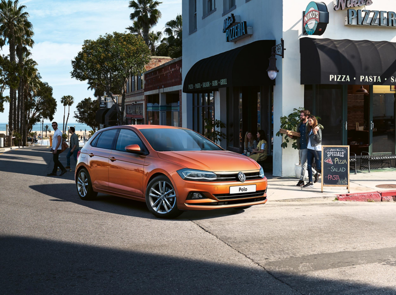 An orange Polo drives past a pizzeria as people walk by