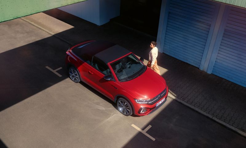 Bird's eye view of the red VW T-Roc Cabriolet with the roof open. Parking on the side of the street, a woman walks past.