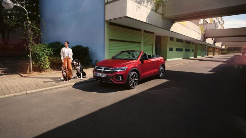 Red VW T-Roc Cabriolet with open roof is parked on the side of the road. A woman with two dogs is standing next to it.