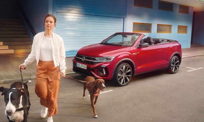A red T-Roc Cabriolet is parked on the side of the road with open roof, a woman with two dogs is crossing the street in front of it.