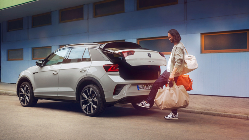 VW T-Roc R-Line in white in front of a building, side visible, man stands behind the car and opens the trunk lid with a swing of his foot