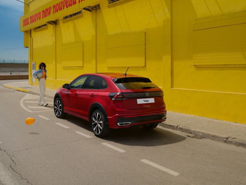 VW Taigo in red on the roadside in front of a yellow building, rear and side view, woman is walking towards the vehicle