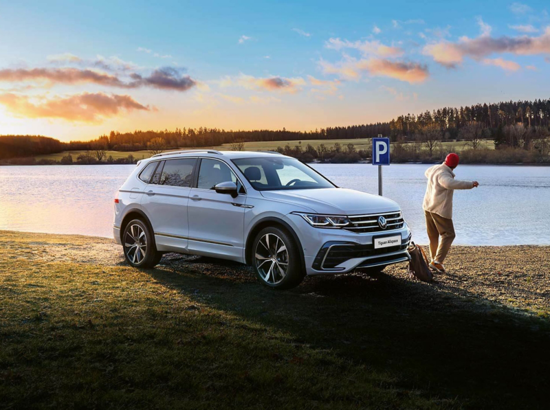 A white Tiguan Allspace R-Line with tinted rear windows and switched-on light signature stands on the lakeshore.