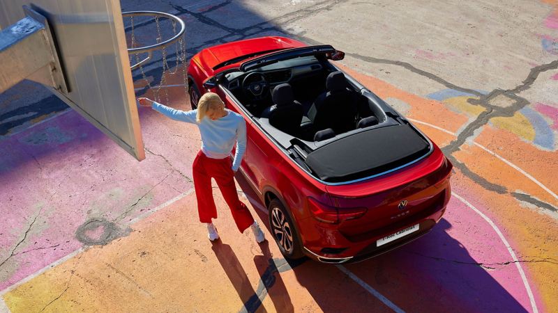 View from above of the red VW T-Roc Cabriolet with an open soft top. A woman is next to it and a basketball basket is visible in the top left corner.