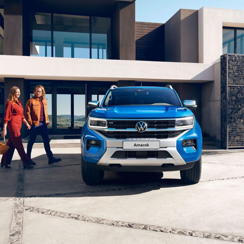 A blue Amarok parked next to a modern house with two people walking towards the car