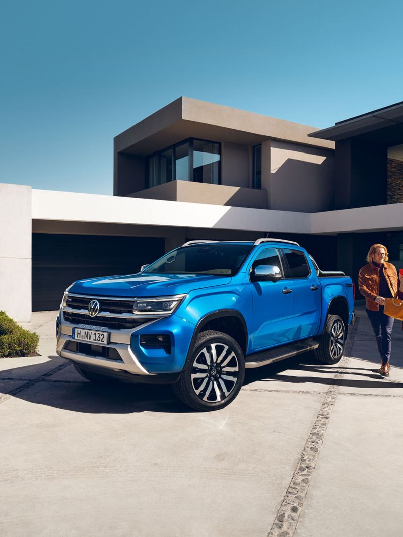 Blue Amarok parked in front of a modern house with a couple walking next to it. 