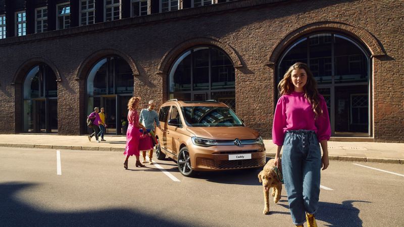 Photo of a woman with a dog walking away from VW Caddy parked in a city. 