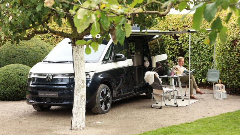 Eine Frau sitzt in einem Campingstuhl und liest ein Buch. Neben ihr parkt ein VW California auf einem Stehplatz im California Garden.