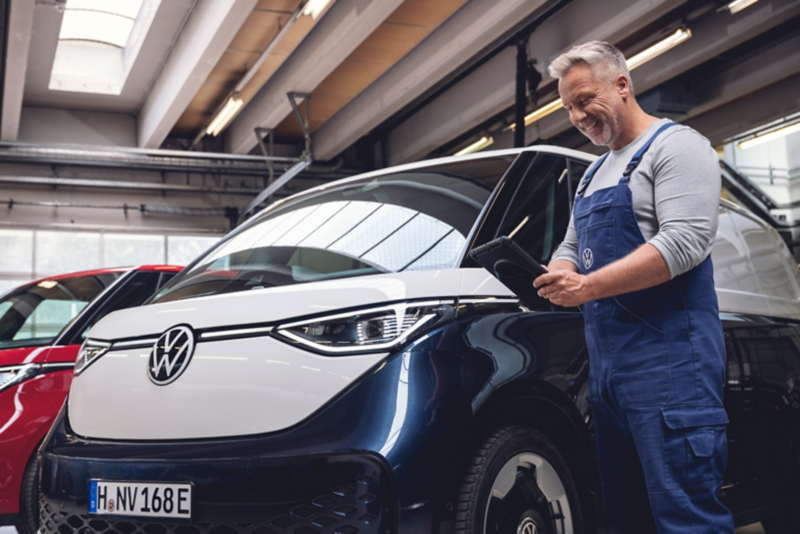 Un artigiano in tuta da lavoro blu si trova in officina accanto a un VW ID. Buzz Cargo e utilizza un tablet per controllare i dati o il software del veicolo.