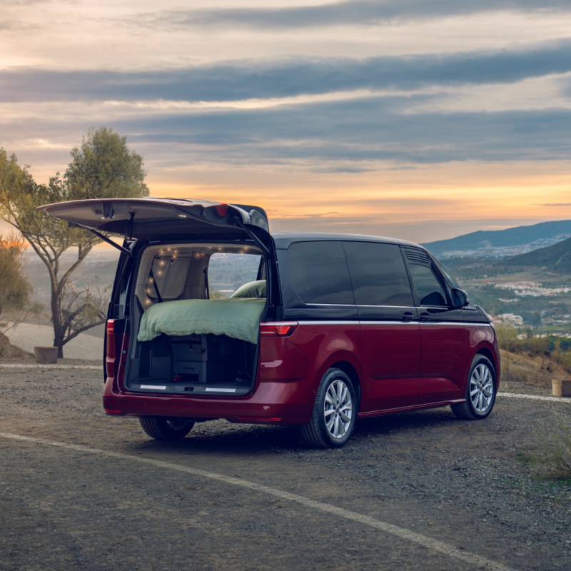 Ein roter VW Van steht auf einem Aussichtspunkt in der Natur. Die Heckklappe ist geöffnet und im Laderaum ist ein Bett aufgebaut, mit Blick auf eine Landschaft bei Sonnenuntergang.