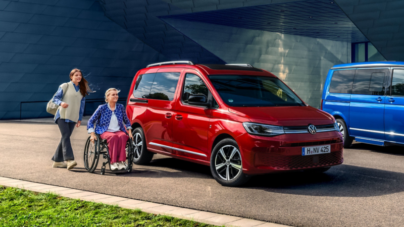 Zwei Personen, eine im Rollstuhl, neben einem VW Caddy vor einem modernen Gebäude