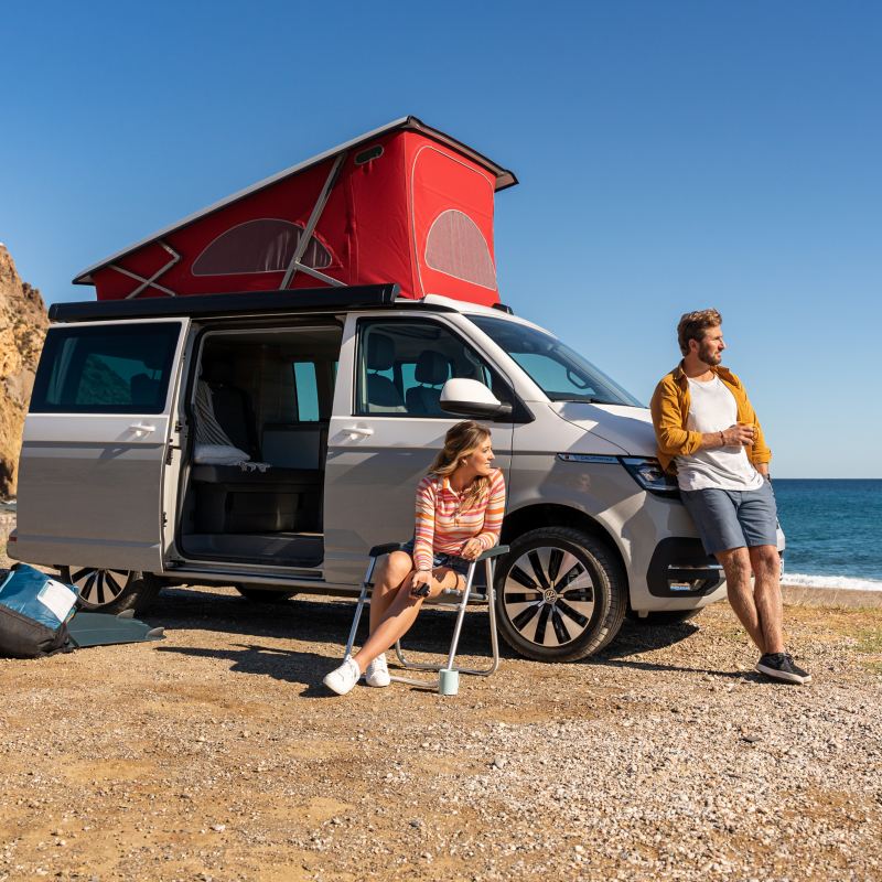 Vw Multivan parkt auf einem Strand am Meer mit ausgeklapptem Dachzelt und geöffneter Seitentür.
