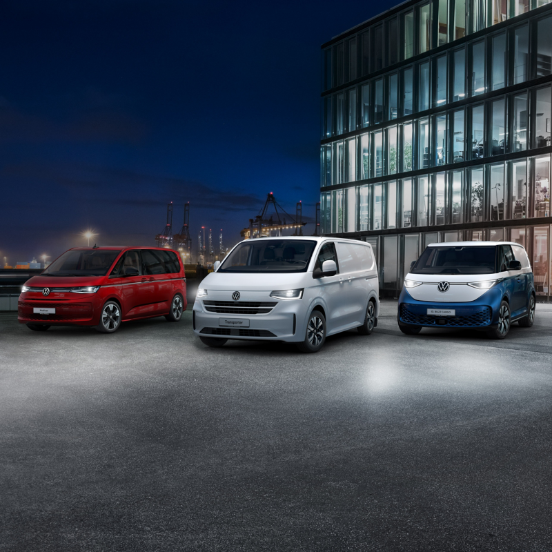 Die elektrisch betriebenen Fahrzeuge von Volkswagen Nutzfahrzeuge parken aufgereiht vor einem modernen Bürogebäude mit Glasfassade. Im Hintergrund stehen Verladekräne eines Hafens bei Nacht.