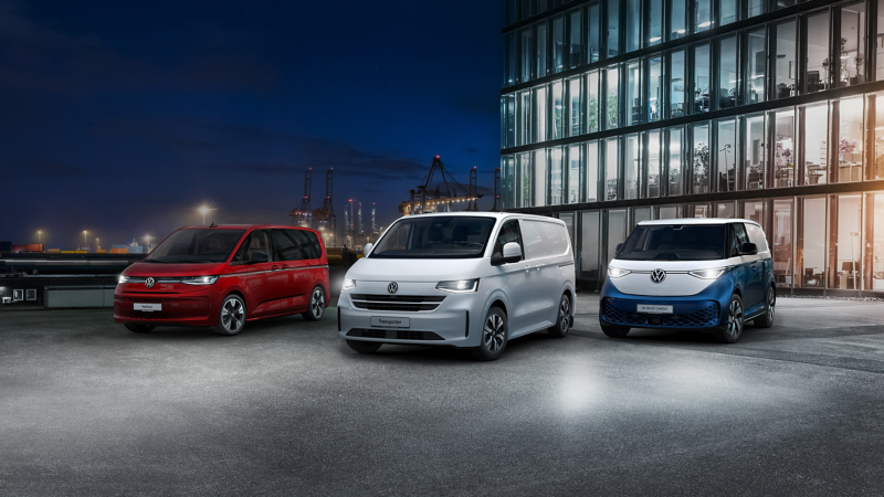 Die elektrisch betriebenen Fahrzeuge von Volkswagen Nutzfahrzeuge parken aufgereiht vor einem modernen Bürogebäude mit Glasfassade. Im Hintergrund stehen Verladekräne eines Hafens bei Nacht.