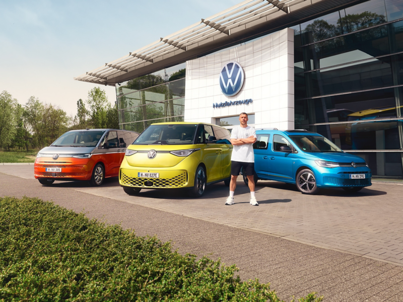 Lukas Podolski steht mit verschränken Armen lässig vor den Volkswagen ENERGY Modellen an einem Autohaus.