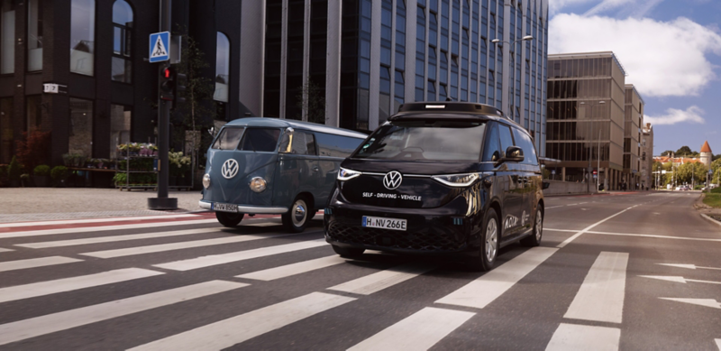 Ein selbstfahrender VW ID. Buzz und ein VW T1 Bulli fahren nebeneinander durch eine Stadt.