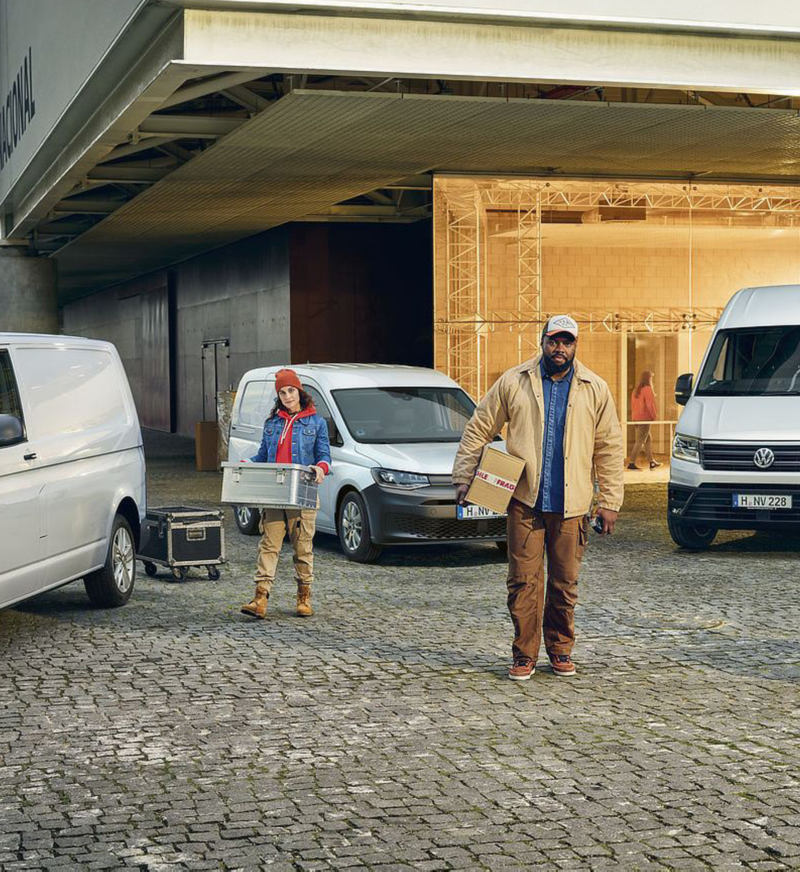 Deux personnes déchargent des colis devant des vans Volkswagen blancs. Un homme porte un colis, et une personne tire un chariot.