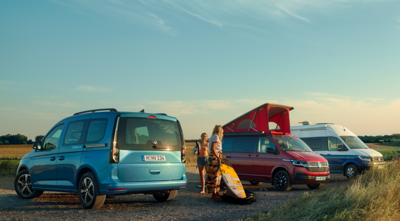 Trois vans Volkswagen (bleu, rouge et blanc/bleu) garés à la campagne au coucher du soleil. Deux personnes déchargent du matériel de sport nautique d'un van rouge.