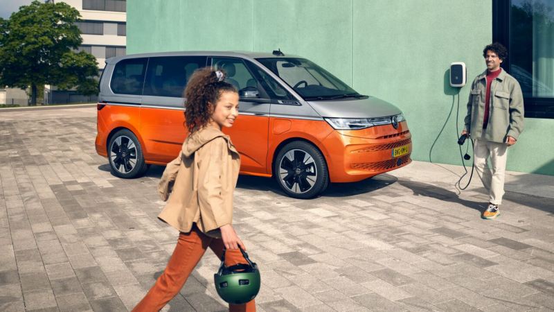 A man with a charging cable in his hand and a girl with a helmet in front of the VW Multivan Energetic.