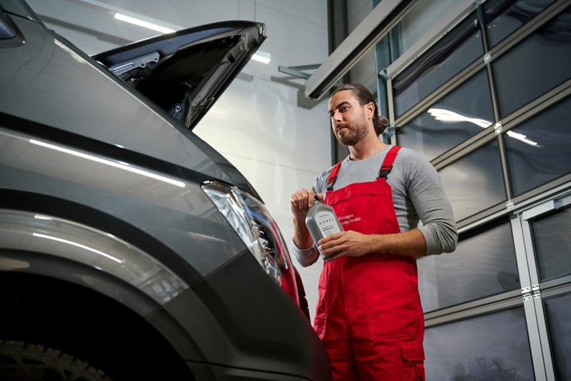 Pracownik serwisu stoi obok dostawczego Volkswagena z otwartą maską, trzymając w rękach butelkę oleju do skrzyni biegów.