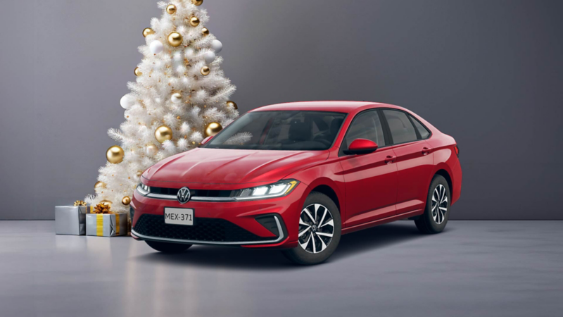 Jetta 2026 en color rojo cerca de un pino de Navidad blanco, decorado con esferas doradas y regalos al pie. 