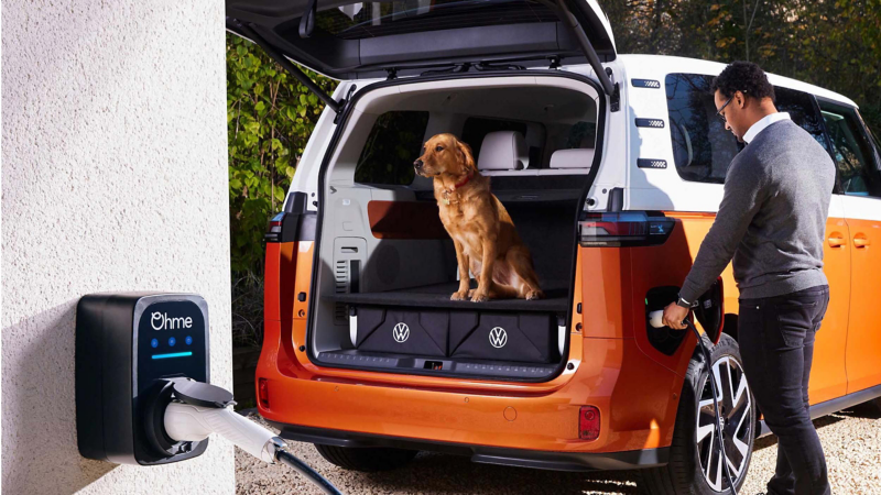 a golden retriever sits in the boot of an orange ID. Buzz while a man plugs it into an ohme home charger